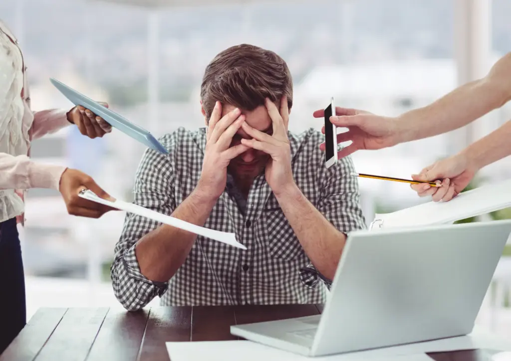 Homem estressado em reunião no escritório com várias pessoas ao redor e ferramentas de trabalho nas mãos, sugerindo sobrecarga e pressão.