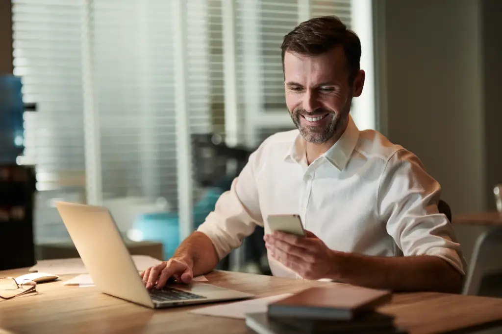 Homem sorridente trabalhando no escritório, usando notebook e celular enquanto lê mensagens em ambiente com luz natural e janelas com persianas