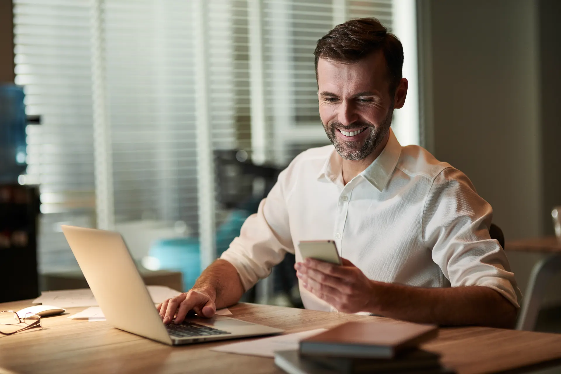 Homem sorridente trabalhando no escritório, usando notebook e celular enquanto lê mensagens em ambiente com luz natural e janelas com persianas